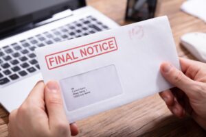 A person holds a final notice letter in while sitting at a desk in front of a silver laptop.