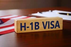 A wooden block that says H1B visa rests on a wooden desk in front of an American flag and paperwork.