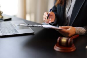Zoomed in picture of a woman sitting at a desk in front of a laptop holding a clipboard and pen next to a gavel