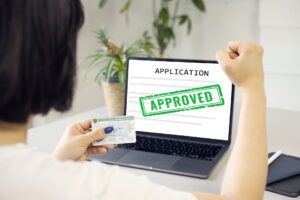 Woman holding a permanent resident card looking at a computer with an application that says approved