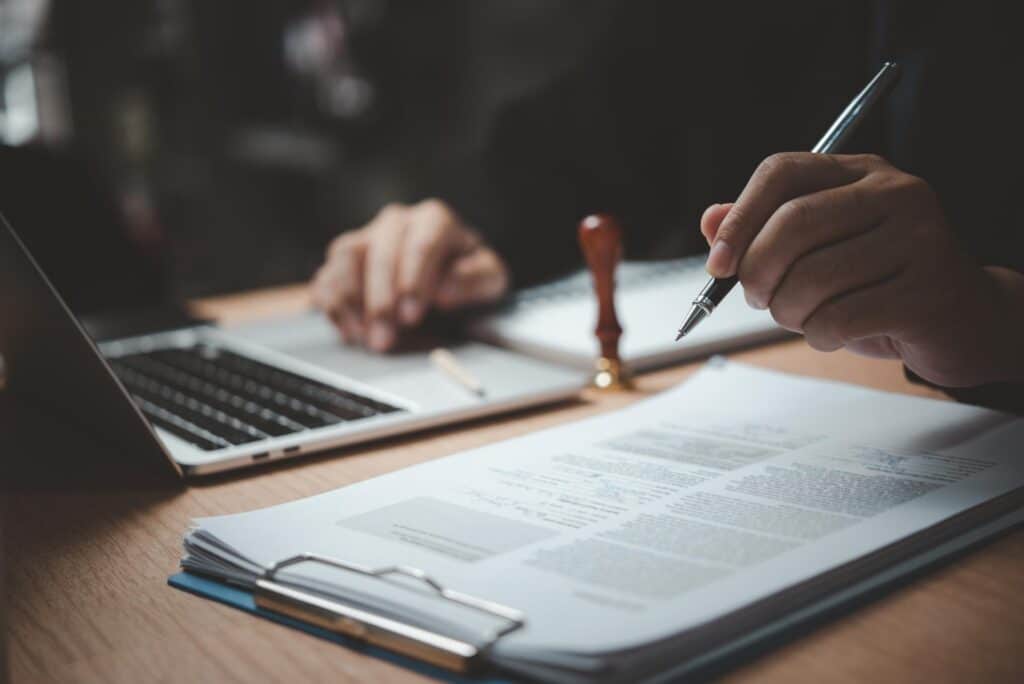 A person holds a fountain pen above a clipboard and paper next to a silver laptop and stamp.