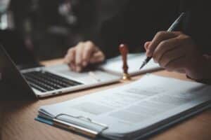 A person holds a fountain pen above a clipboard and paper next to a silver laptop and stamp.