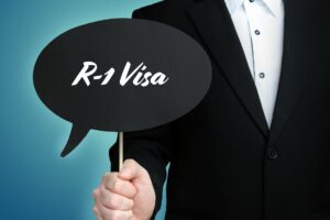 Person in a black suit zoomed in on and holding a sign that says R-1 Visa