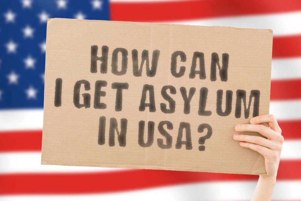 A sign being held up that says "How can I get asylum in USA?" on top of an American flag
