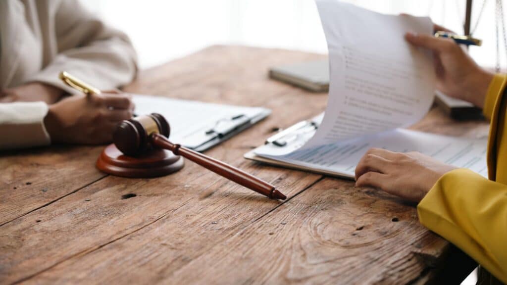 Two people sit at a desk reviewing paperwork on clipboards next to a gavel resting on its stand.